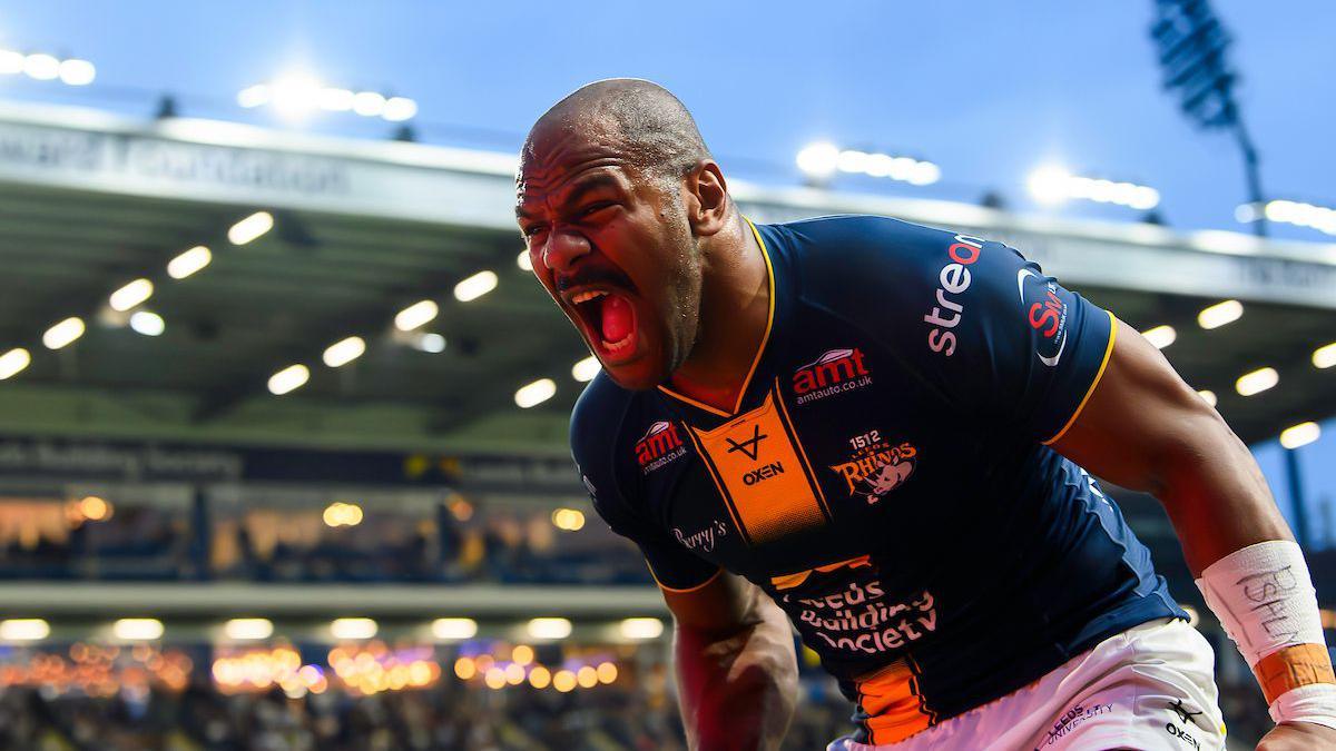 Leeds Rhinos wing Maika Sivo, in a blue and amber shirt, shouts in celebration of a try, with a Headingley stand and floodlights in the background.