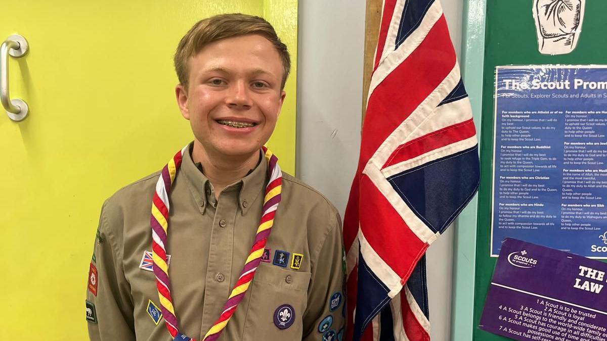 A teenager wearing a brown Scout uniform shirt and neckerchief, in front of a Union flag.