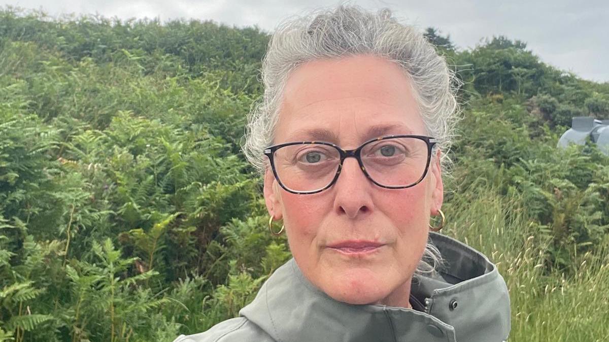 Emily Crichton looking directly wat the camera. She has tied-back grey hair and is wearing glasses, and is standing in front of a bank of foliage.