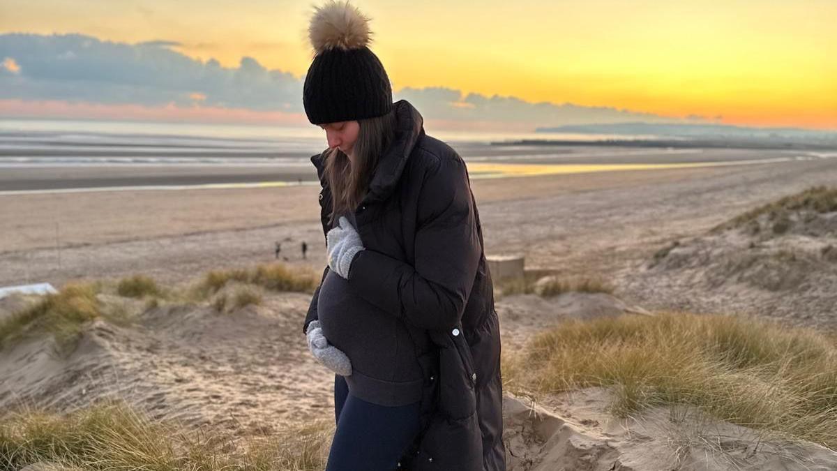 Samantha during pregnancy standing in sand dunes by the sea, holding her stomach and looking down at it