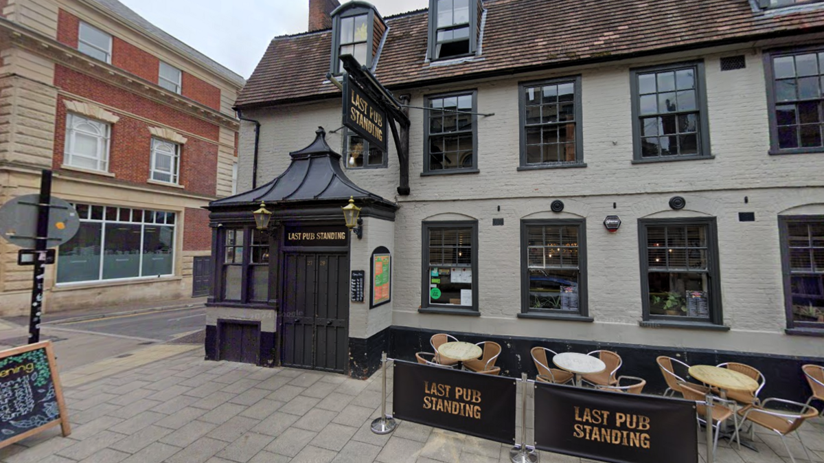 A pub with grey walls and dark grey window and door frames is shown with its name signs above the door, with an A-board advertising its events to the left and and an empty seating area to the right.