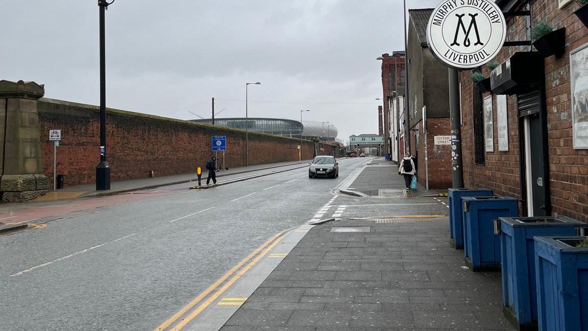 A view down Regent Road with Everton Stadium in the distance, one side of the road is lined with a large brick wall and the other with former dockland buildings