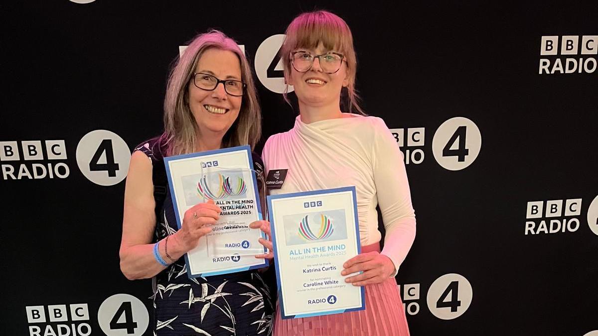 Two women are standing side by side holding up certificates and a glass award. They are both smiling. They are in front of a black back drop with the BBC Radio 4 logo printed on it.