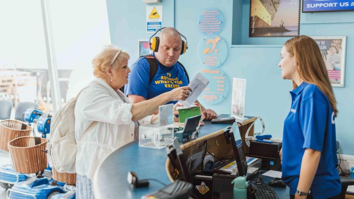 A bald man with yellow headphones on and a blue top that says 'I'm autistic what's your excuse' is stood at a desk with a woman next to him with short blonde hair and a white outfit who is holding up a piece of paper to him. A young woman in a blue polo shirt with long brown hair is stood behind the desk in front of a computer.