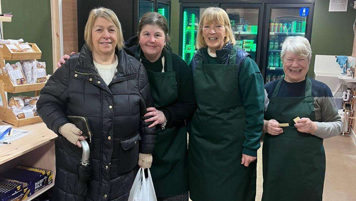 Three shop volunteers in green aprons stand alongside their first customer dressed in a black coat