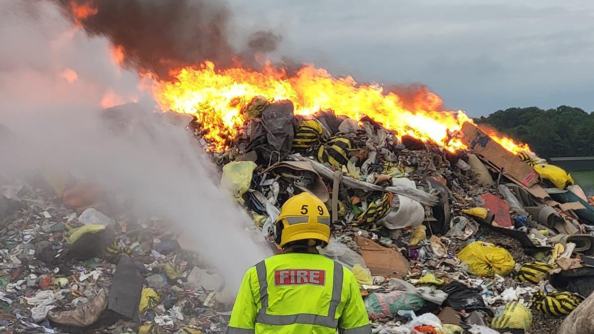 Firefighter with water hose spraying piles of rubbish with fire in background