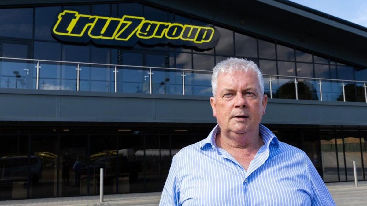 A photo of Guy Nichols who is wearing a light blue shirt and standing in front of a large black and glass building with a yellow and black sign which reads tru7group