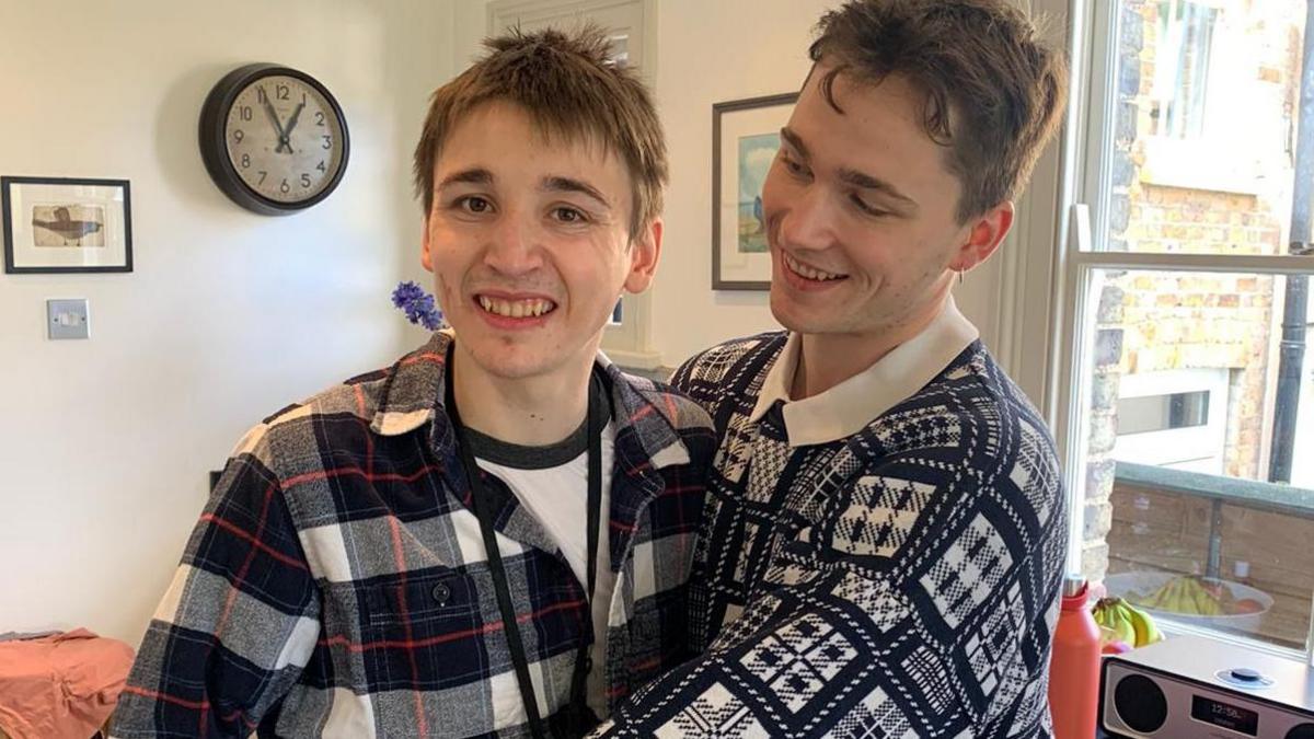 (l-r) Felix Geldart is smiling at the camera has a blue and white checked shirt on and his brother Jude is hugging him and looking at him.