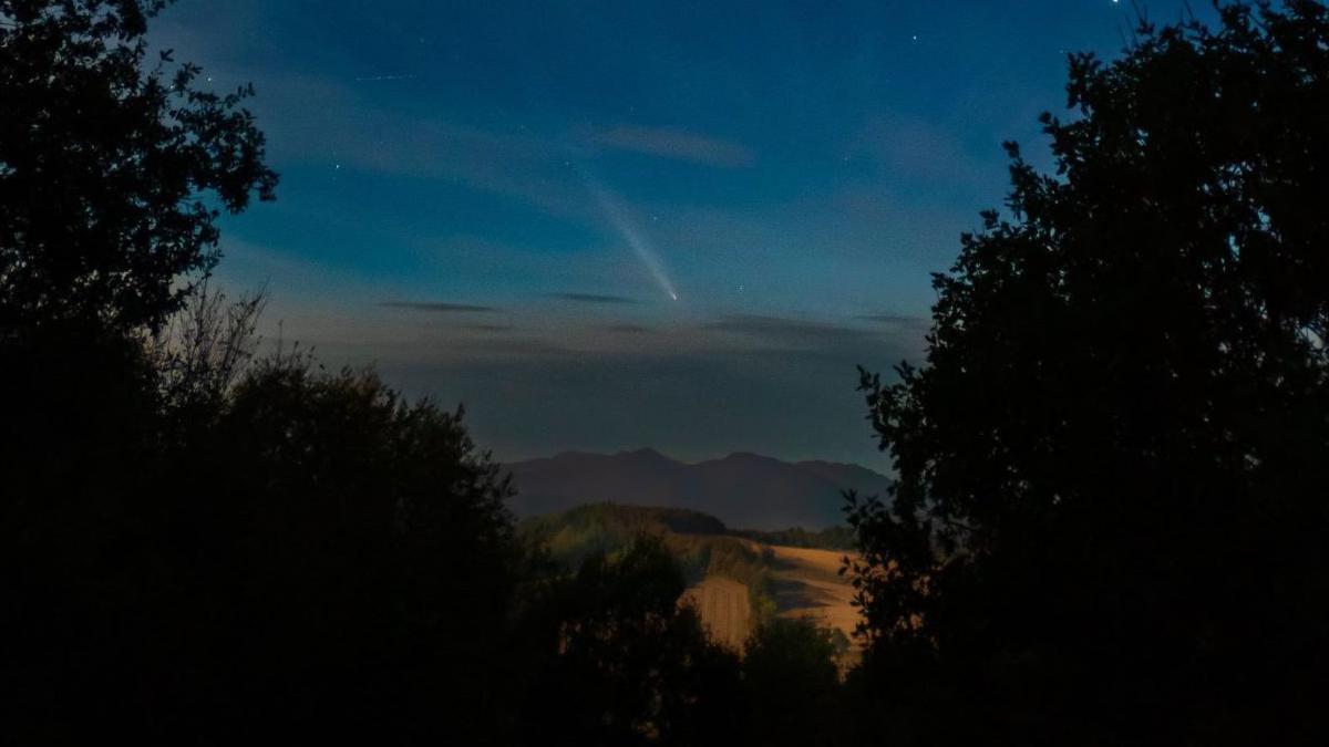 Comet A3 appearing above hills and fields and framed by trees.