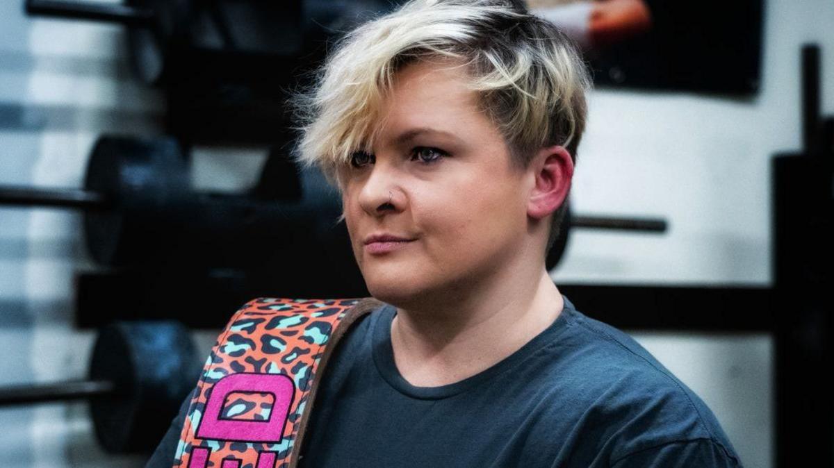 Andrea Seed who has short blond hair and green eyes and nose ring wearing a black t-shirt with weights in the background with an orange blue and black weightlifting belt which has her surname 'Seed' on it, hanging on her shoulder