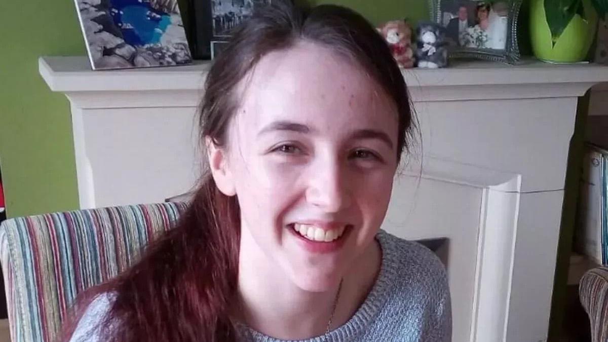 Llun o Catrin Maguire yn gwenu at y camera wrth eistedd yn ei ystafell fyw yn gwisgo siwmper llwyd