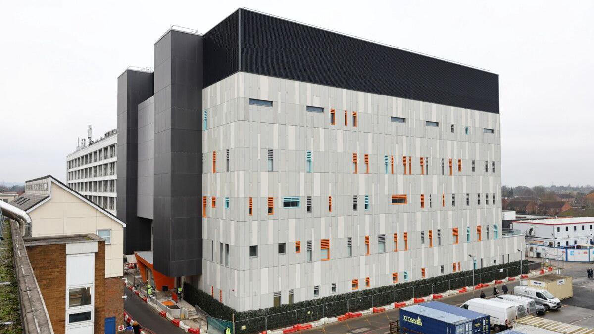A large new block at a hospital which is at least five storeys high. There are vans and lorry containers in the foreground.