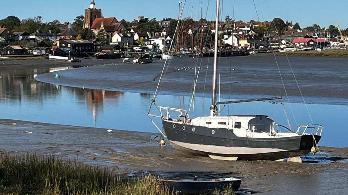 A white and navy (or black) boat sitting in mud at Hythe Quay. Water and mud stretches into the background and a collection of houses, including St Mary's church, can be seen in the background