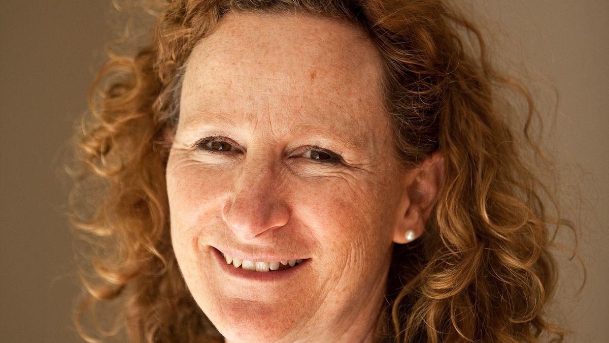 Jane O'Riordan smiles at the camera. Her head is turned at a slight angle to look at the camera. She has curly red hair and has pearl earrings.