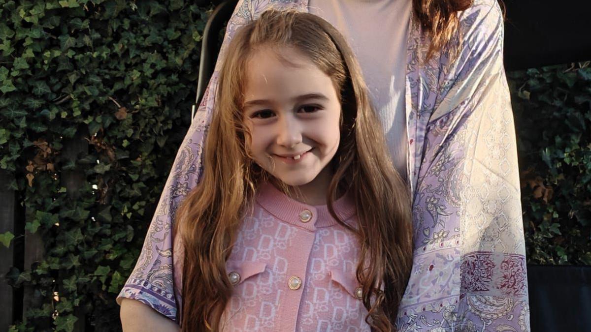 A girl with long brown hair is standing in a garden. The child is wearing a pink cardigan with a geometric pattern and blue jeans. A woman is standing behind the girl. She is wearing a long patterned kimono-style garment over a beige top. Behind them is a fence covered in green ivy.