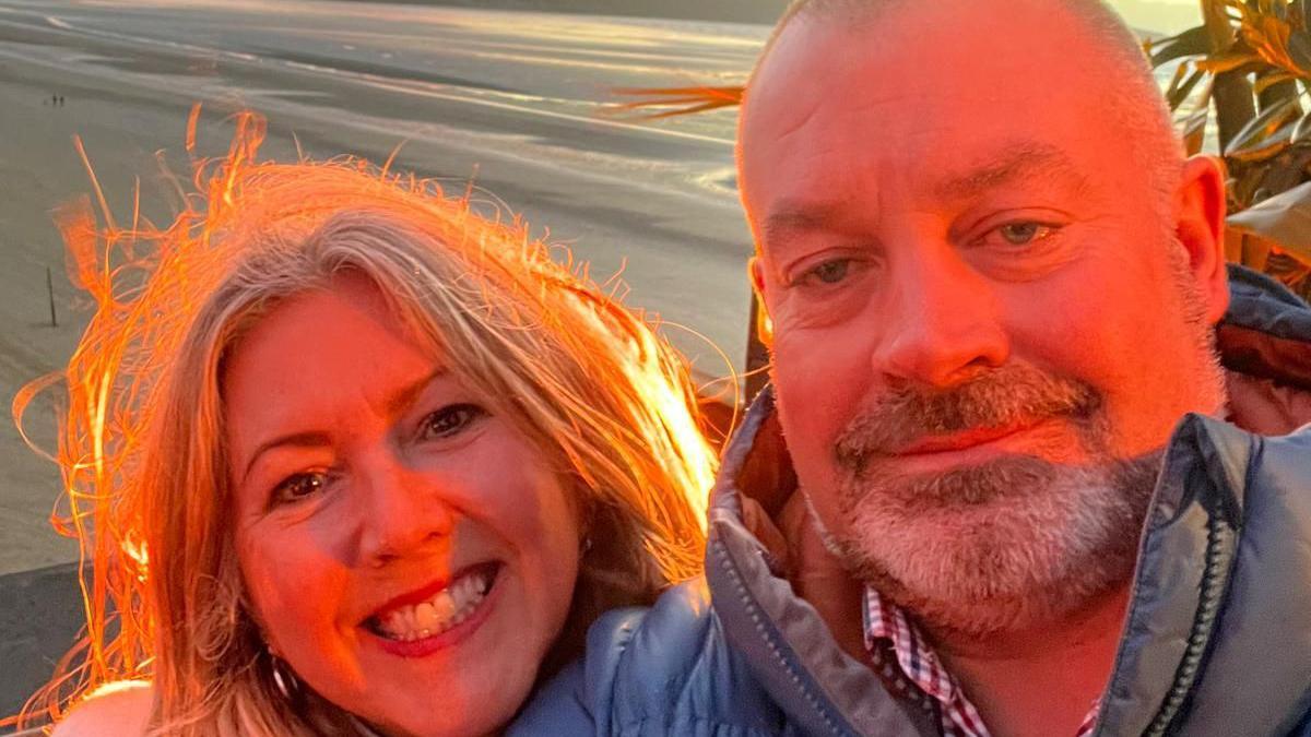 Joanne Brandon-Hodgkinson with David Griffiths. Joanne has shoulder length blonde hair. Griffiths has cropped grey hair and a beard, He is wearing a checked shirt and a puffer jacket. They are smiling and by the sea.