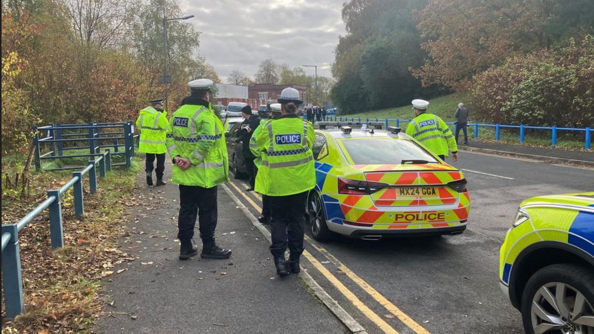 Four police officer surround a black car on foot on a road outside HMP Forest Bank on an autumn day, with two police vehicles parked behind.