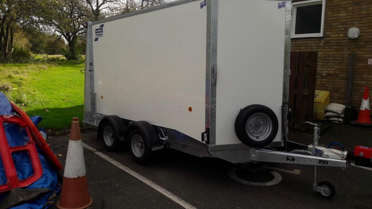 A grey trailer in a parking bay. The trailer is metal and not attached to a vehicle. To its right are parking cones and discarded road signs.