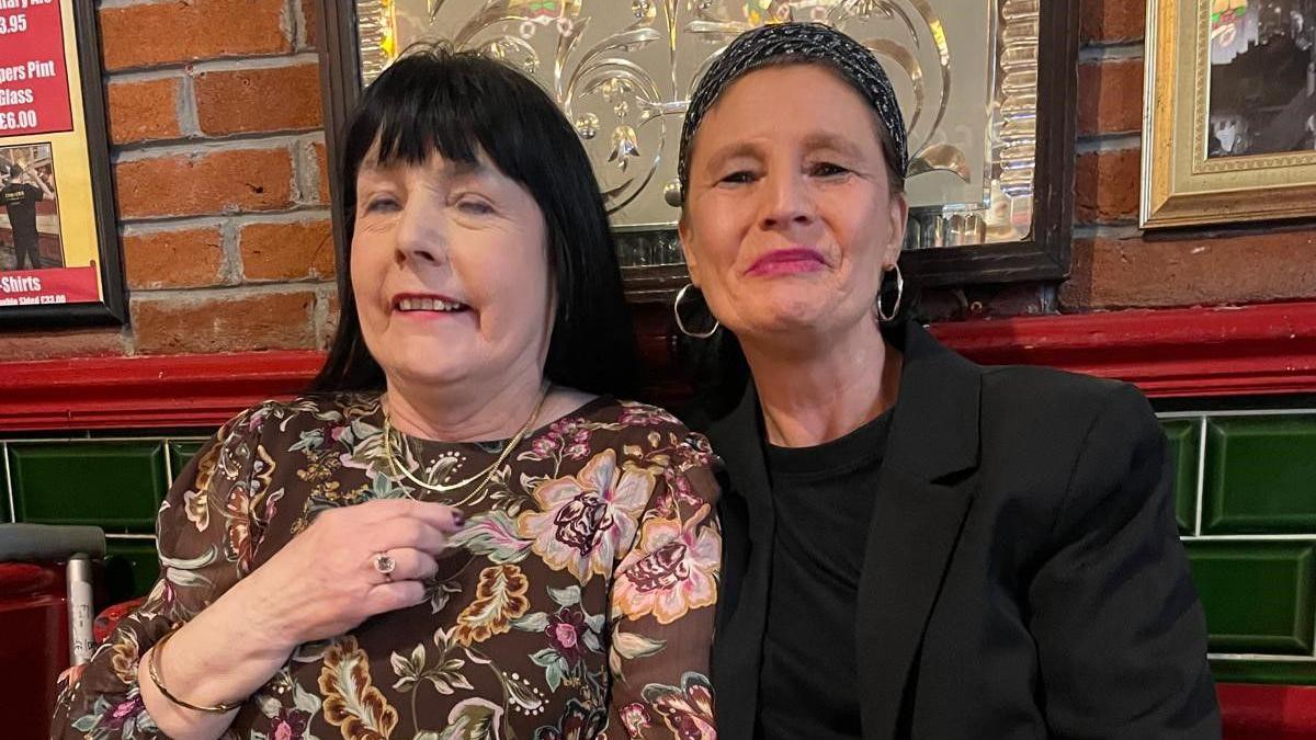 Two women sat in the pub. One has mid length black hair and is wearing a brown flowered top, the other has a head band on and has hoop earrings and is wearing a black t-shirt and black jacket. 