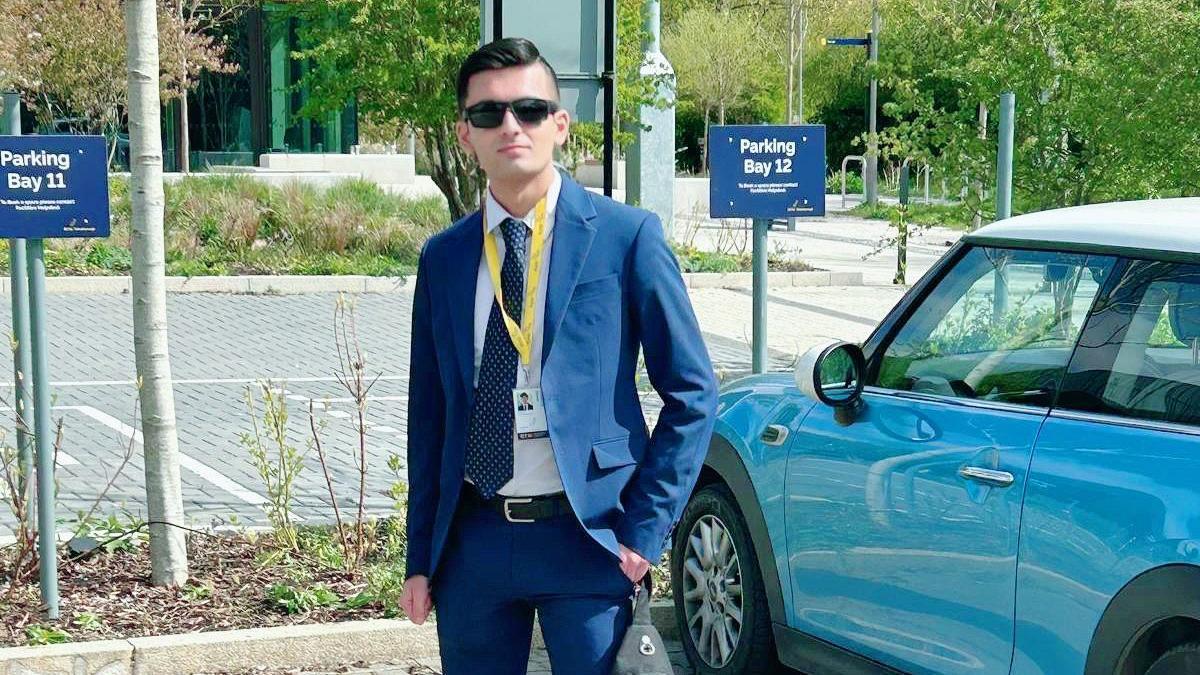 A young man in a navy suit with a white shirt and blue and white spotted tie is standing next to a blue Mini car in a car park. He has short brown hair slicked back a bit and is wearing black sunglasses.