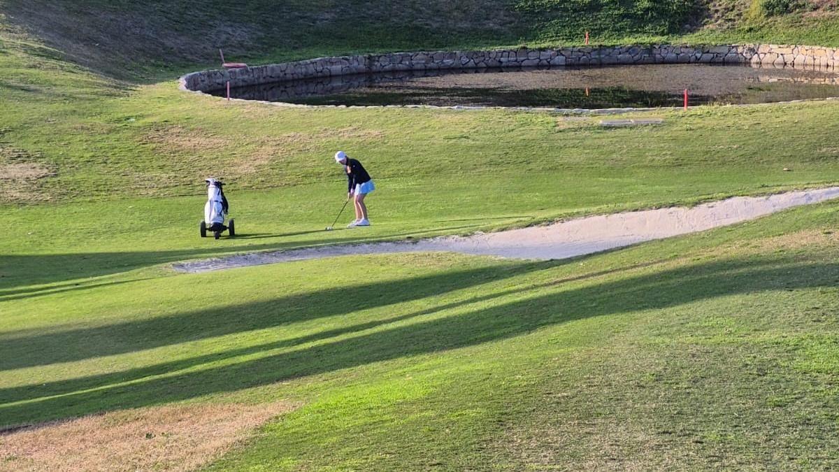 A young girl in the distance gets ready to play a golf shot towards the camera from near a sand bunker on a golf course. She is wearing a white cap, a dark top and a white skirt or shorts. Near her is a wheeled golf bag.