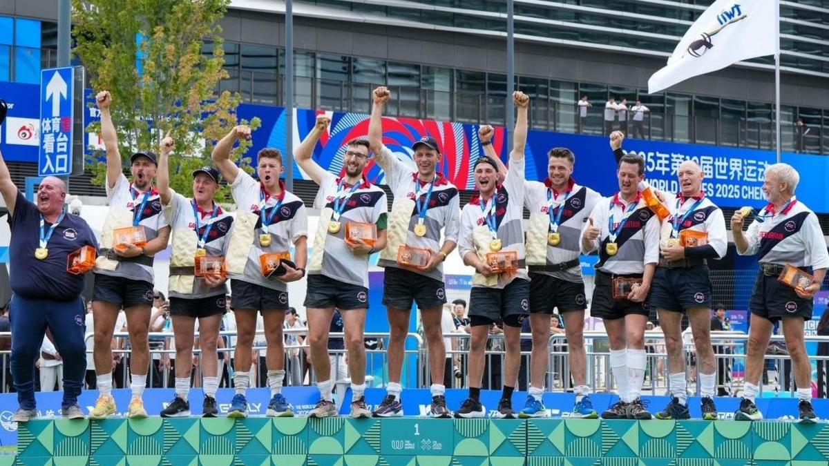 Joe Birch and his team members on a podium wearing their medals - all or their arms are held up in victory. 