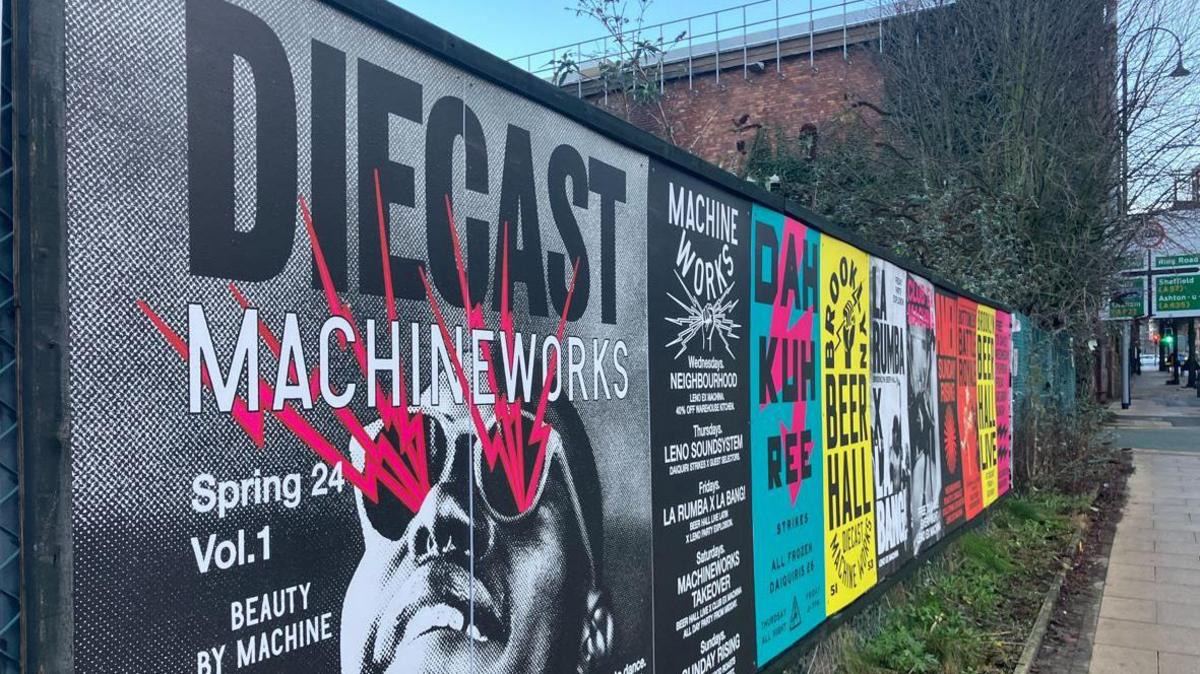 A display board running alongside a pavement with colourful posters showcasing the events being held at the venue. 