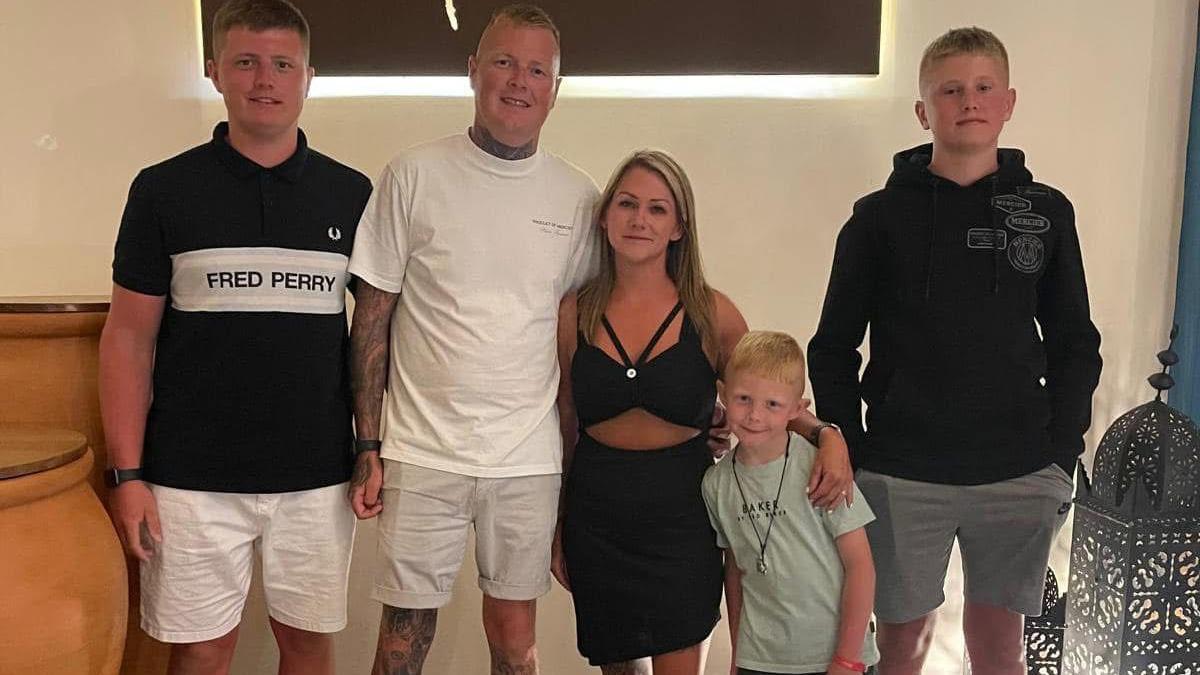 Koby Jones (far left) pictured with his family. He is wearing white shorts and a T-shirt printed with the words Fred Perry. He is stood next to a man wearing a white T-shirt and light shorts. Also stood in a line is a woman, wearing a black dress with her arm around a young child. There is a teenage boy stood at the end of the line wearing grey shorts and a black hoodie.