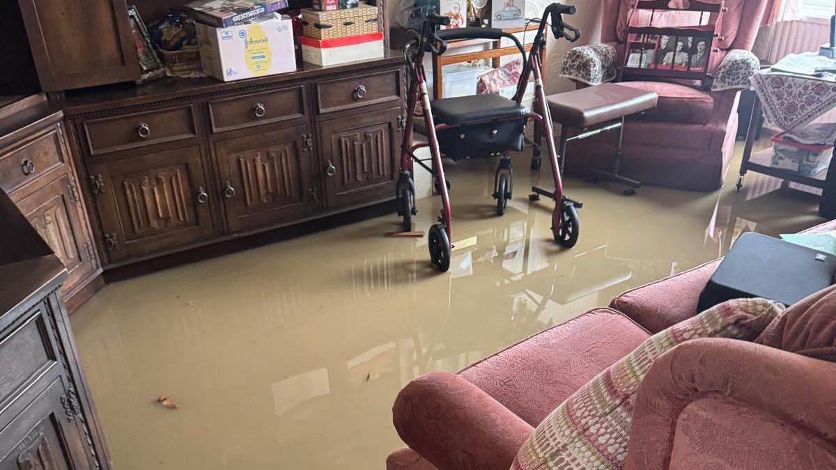 A living room, with a red sofa and a walking frame, with flood water covering the floor. 