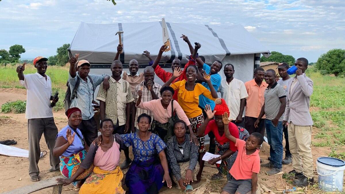 A large group of people stand together with their arms in the air. Some are smiling and offering a thumbs up to the camera, some are holding tools. They are dressed in a variety of bright clothing. There is a tent behind them. 