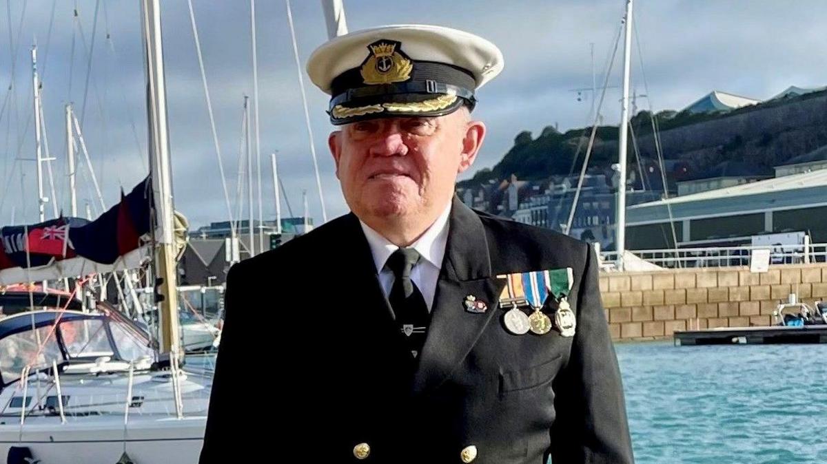 Captain Stanley Richard-dit-Leschery wearing his Royal Naval Reservist uniform with three medals stands in front of the harbour looking at the camera