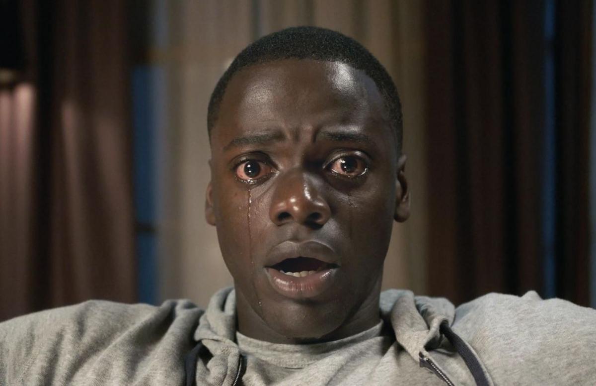 A scene from the film Get Out. Chris Washington, played by Daniel Kaluuya, seated and dressed in a grey hoodie and t-shirt, has a shocked and scared expression as tears roll down his face. 