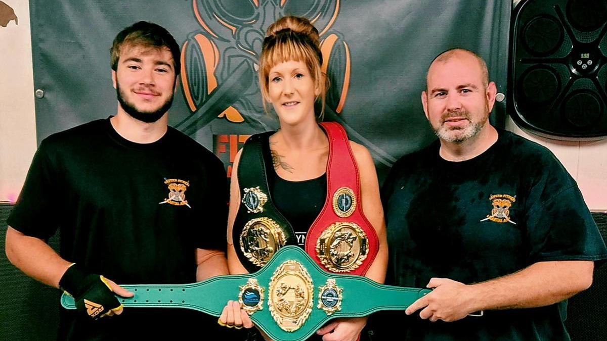 Lucy Payne holding teal belt with two men stood either side. 