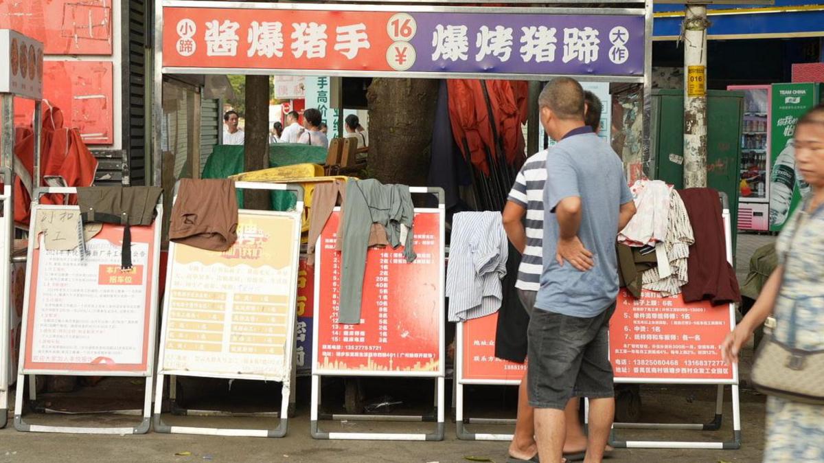 A man in a t-shirt and shorts looks a row of red and yellow bulletin boards with job ads on them and clothes draped over them.