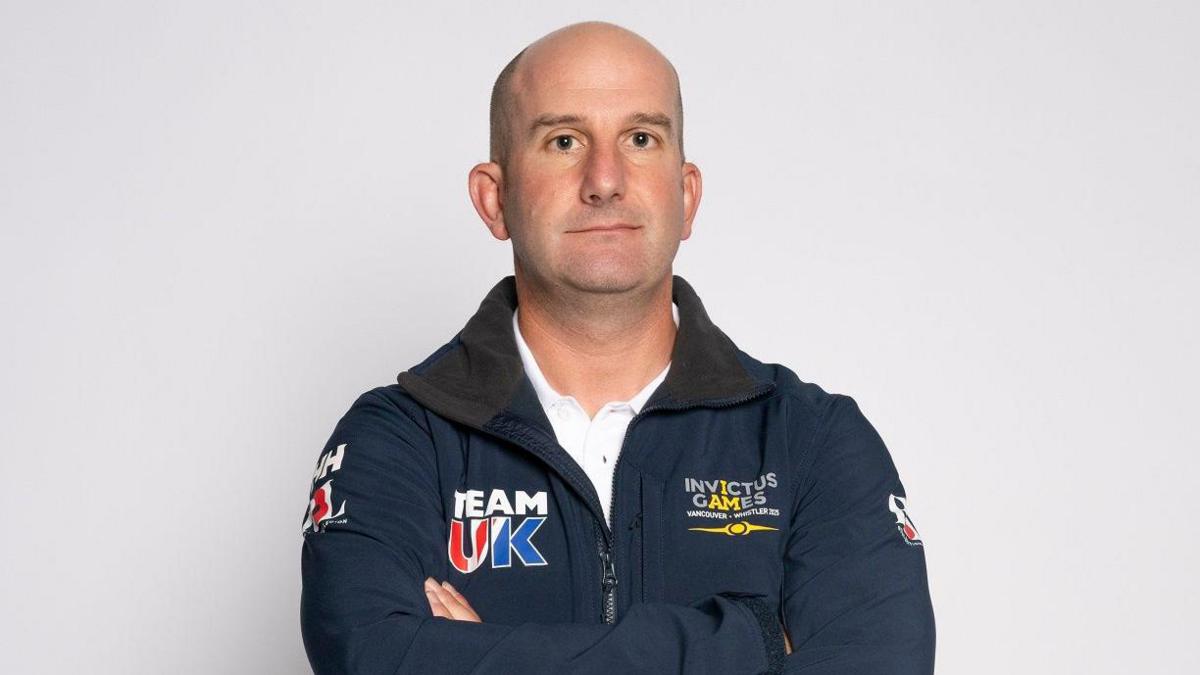 A bald white man in a navy blue branded jacket and a white shirt stands in front of a blank backdrop.