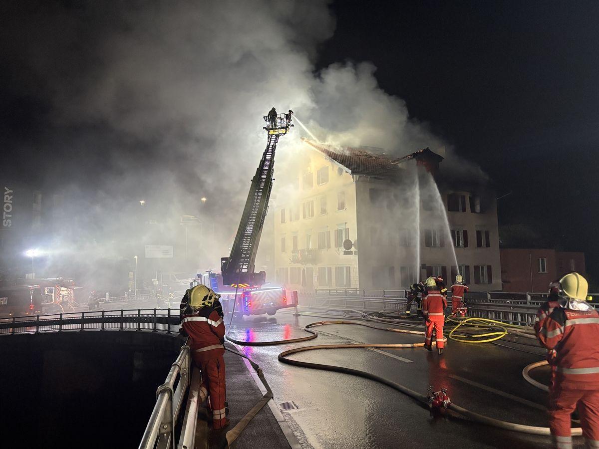 The building as seen from a bridge crossing a river. Three streams of water from firefighting crews douse the building. Smoke rises from the building - at night.