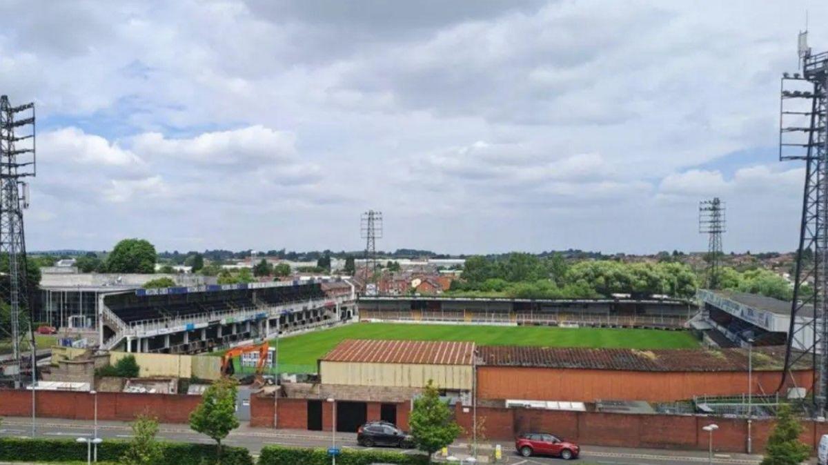A football stadium, with the pitch in the middle surrounded by stands. In each corner of the stadium is a tall floodlight structure.
