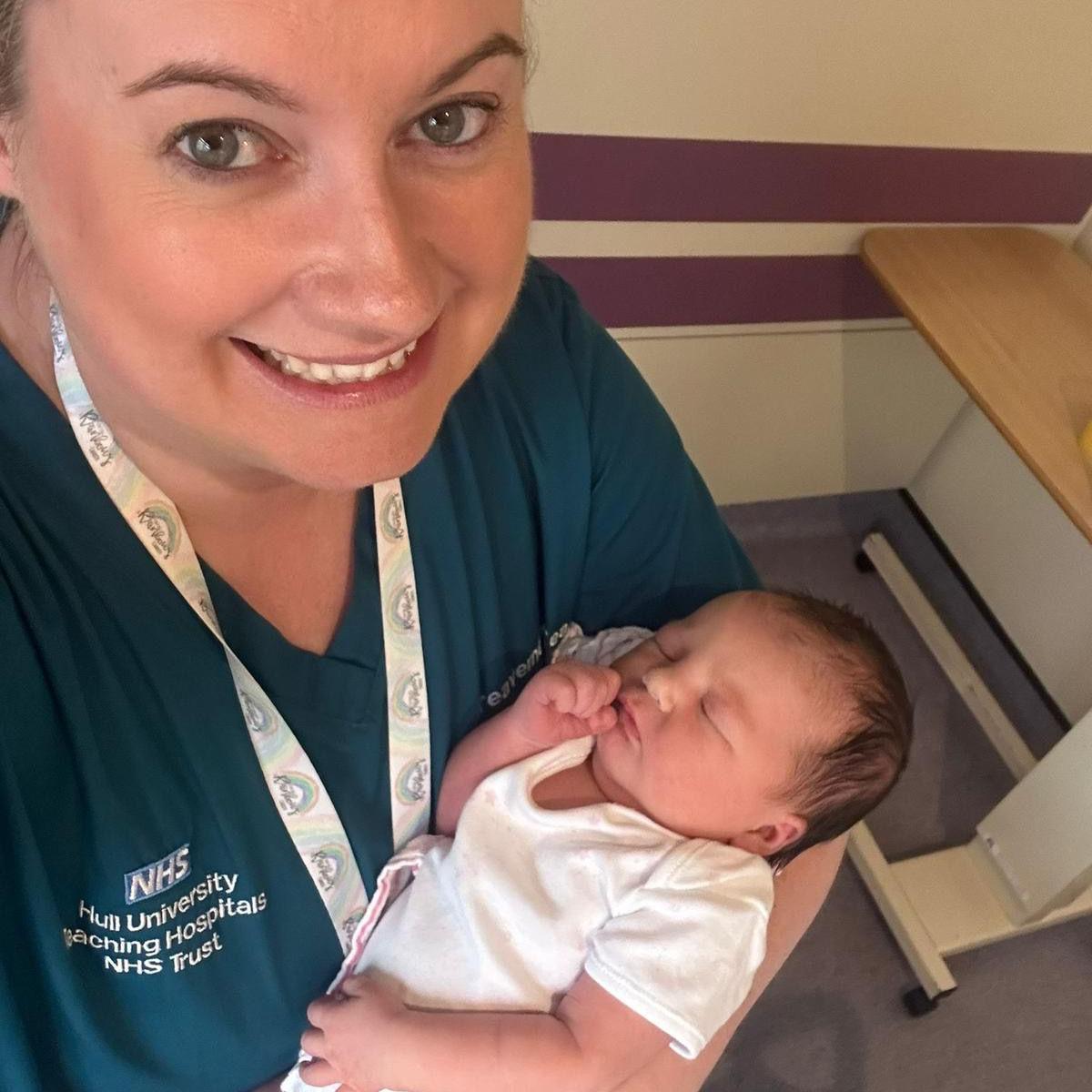 Sam Catanach, who wears a white lanyard and dark-blue scrubs with an NHS logo, smiles as she holds a newborn baby, who has short, dark hair and wears a white top. 