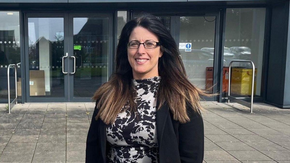 Menyw gyda gwallt hir tywyll syth a sbectol yn gwisgo siaced ddu a ffrog ddu a gwyn. Mae'n sefyll o flaen adeilad ysgol gyda'r gair 'Croeso' arno.