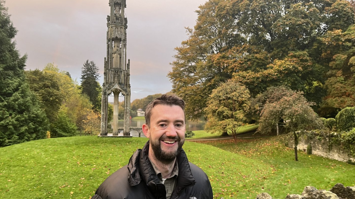 Tim Parker smiles into the camera, wearing a black North Face puffer jacket, with the Stourhead gardens behind him