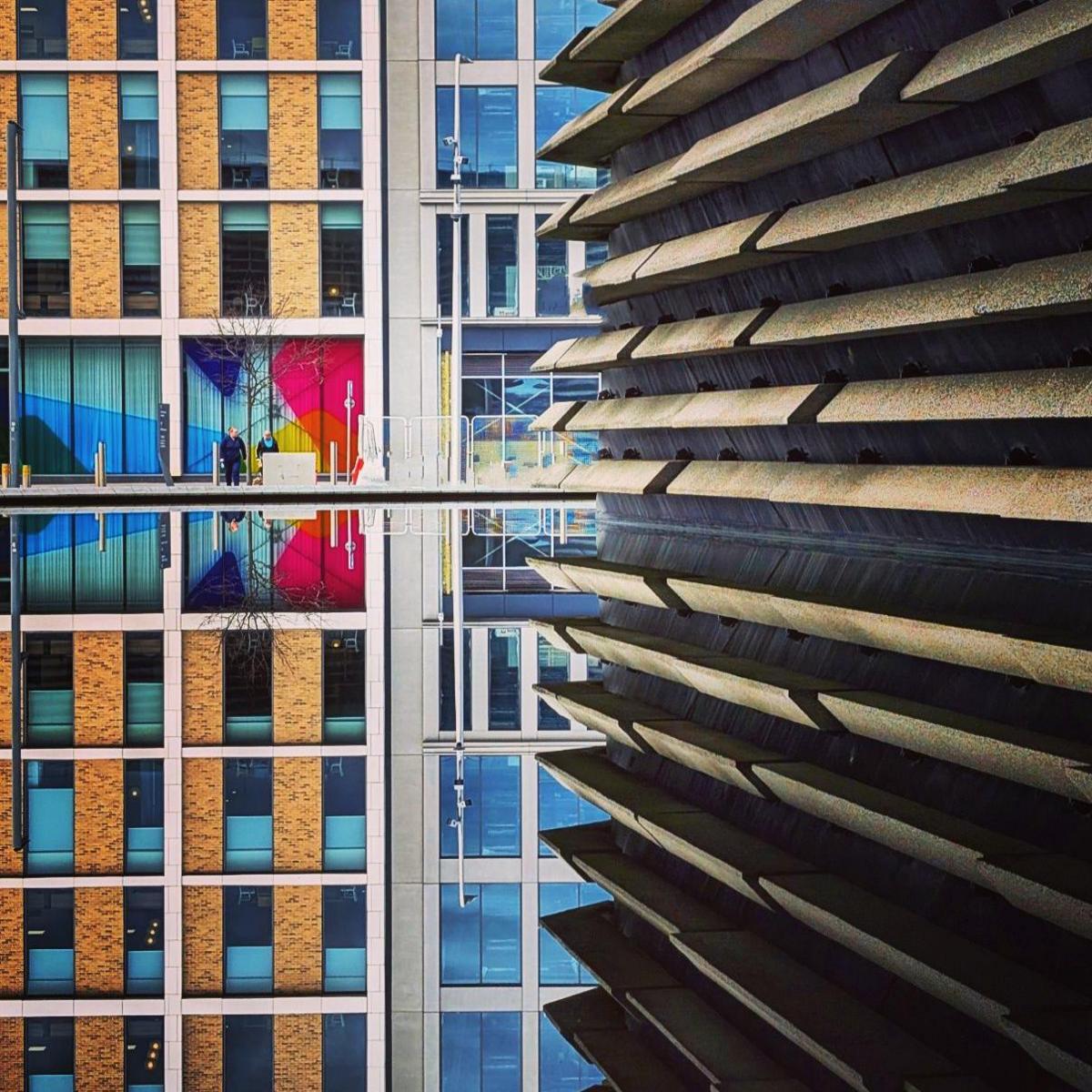 The side of the V&A building in Dundee which is slatted is on the right of the image. It is reflected in the water at the bottom of the picture. Two other buildings can be seen in the background.