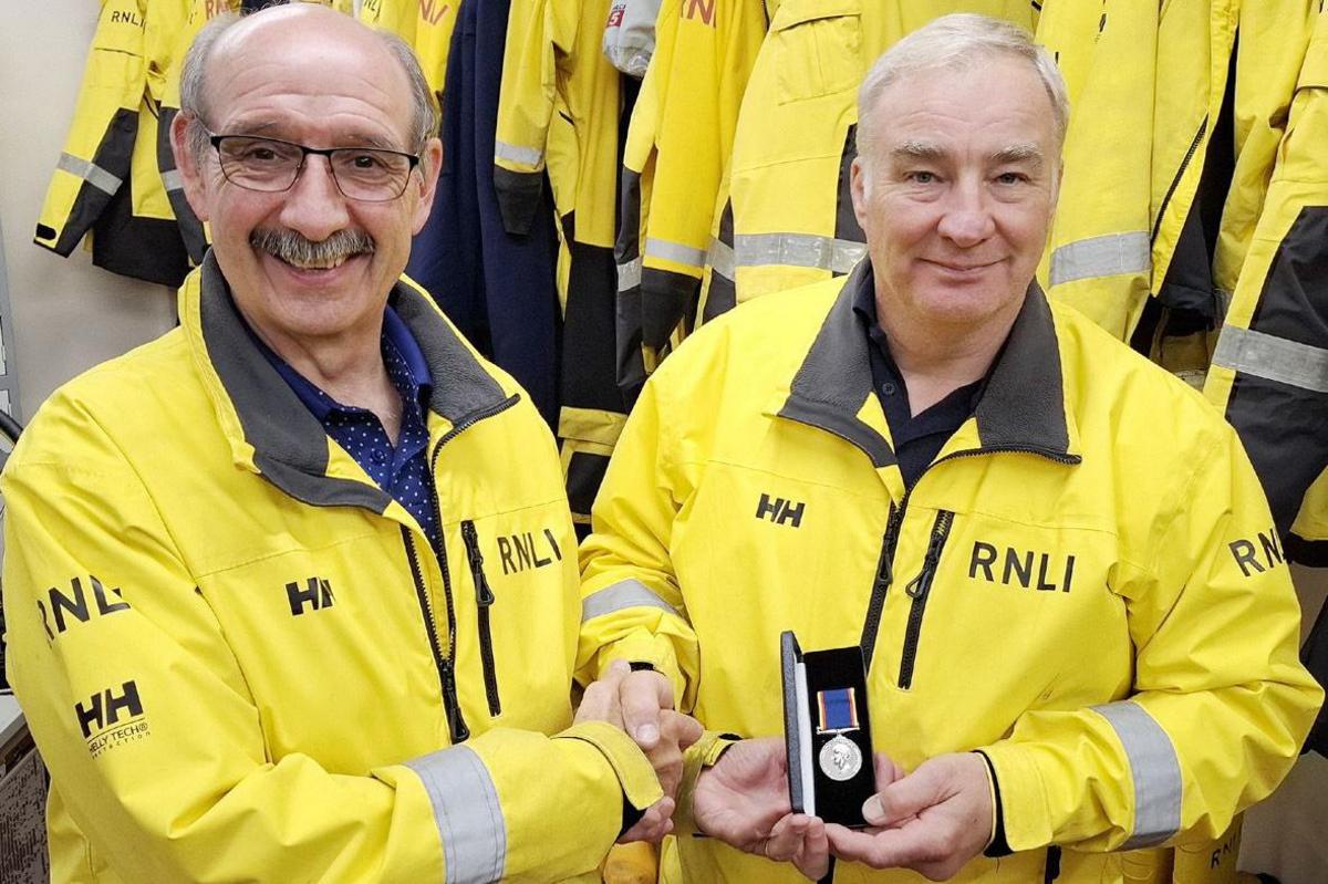 Steve Pounder smiles as he presents Mark Barker with his medal in a box. They are both wearing yellow RNLI jackets.