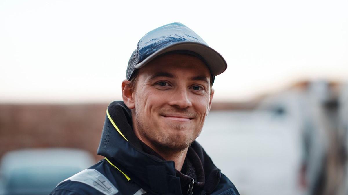 Freddie Watson is a man with a blonde beard. He is smiling into the camera. He is wearing a blue cap and a navy waterproof with yellow trim. The background is blurred out behind him.