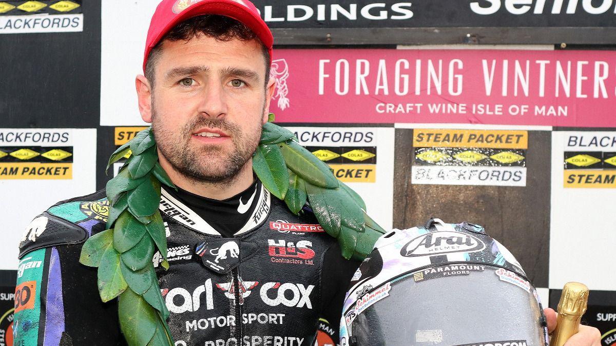 Michael Dunlop in black, turquoise and purple leathers with Carl Cox Motorsport written across the chest. He's wearing a pick peak cap and holding a white helmet featuring the same colours as his leathers. He also has a green garland around his neck and a champagne bottle in his hand. There is a board featuring sponsors' logo in white, black, yellow and pink behind him.