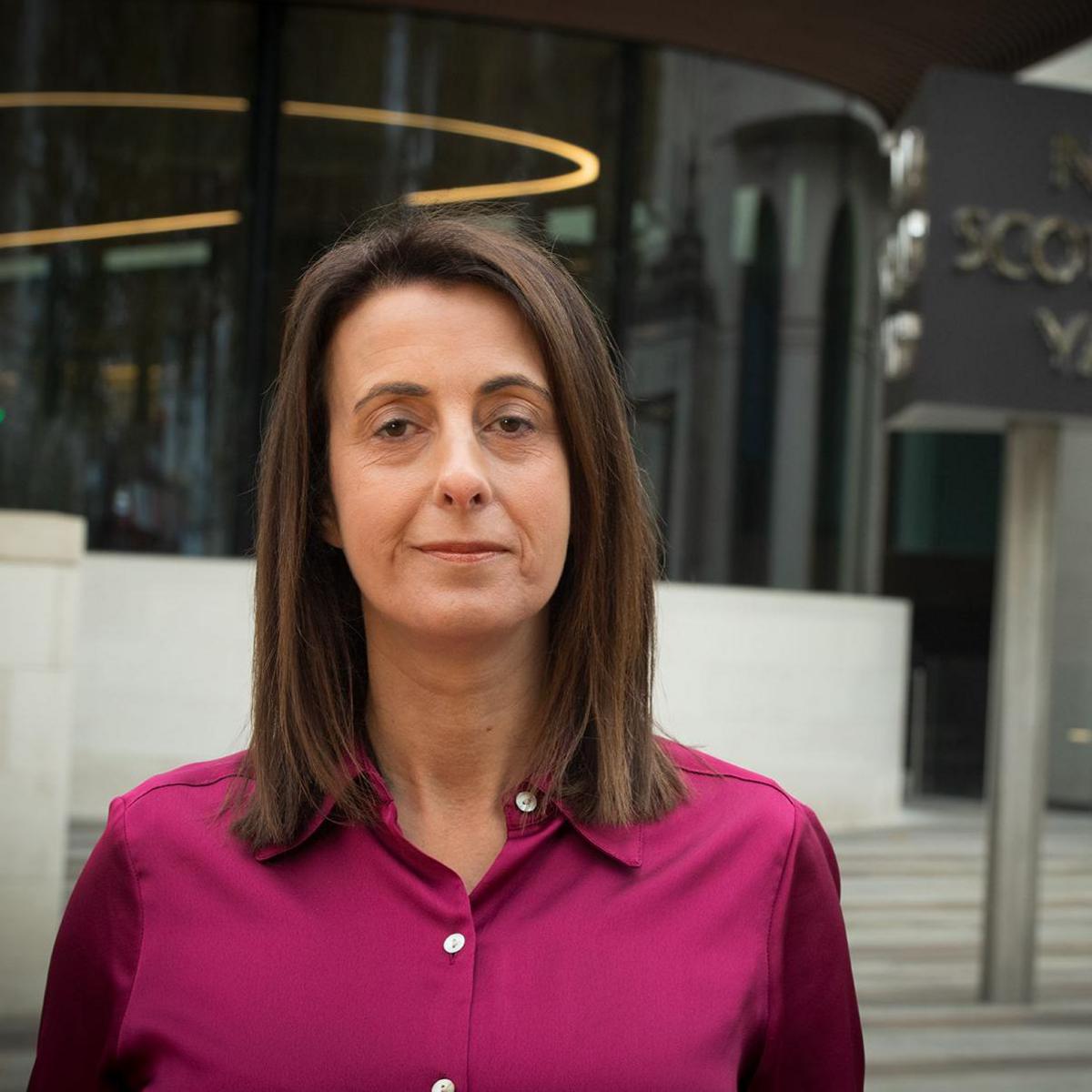 Lucy Manning, a woman in a pink blouse with brown hair, stands in front of a building looking at the camera. There is a sign that says "New Scotland Yard".