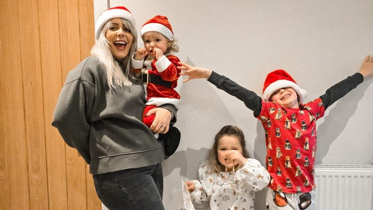 Katie Tradie with her three children, her youngest child sits on her hip in a full red and white santa costume, while her son and daughter stand to the right of her. Katie has blonde mid length hair, she wears a grey sweatshirt and a red santa hat. 