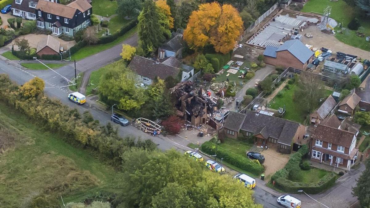 A drone shot of a destroyed detached house close to other large houses that are untouched. Police tape and emergency vehicles fill the road outside.