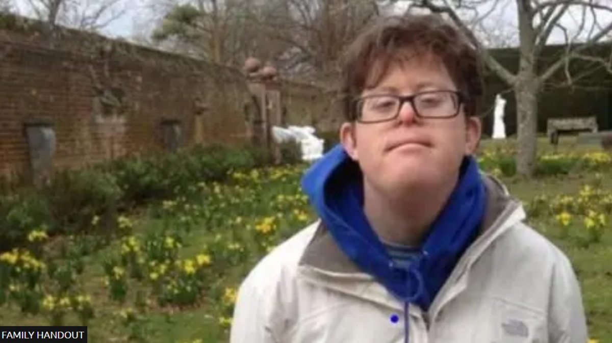 A man with glasses stands in a grassy area surrounded by yellow flowers. He is wearing a blue hoodie under a white coat and has long-ish brown hair. 