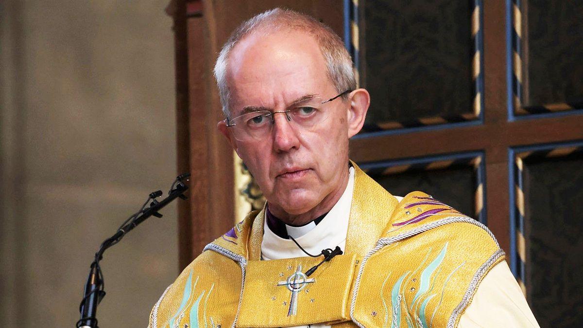 Archbishop of Canterbury Justin Welby pictured in front of a small microphone in a church pulpit. He has receding short grey hair, rimless glasses and is clean shaven. He is wearing gold coloured robes embellished with purple and turquoise waves and jewels, and a silver embroidered crucifix. 