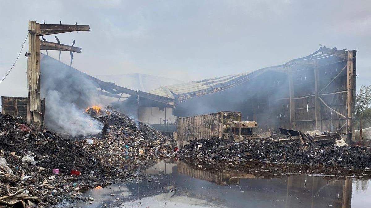 A building destroyed by a fire - its walls are close to collapse and there are mounds of black debris on the ground. The remains of a burned-out truck is on the right. 